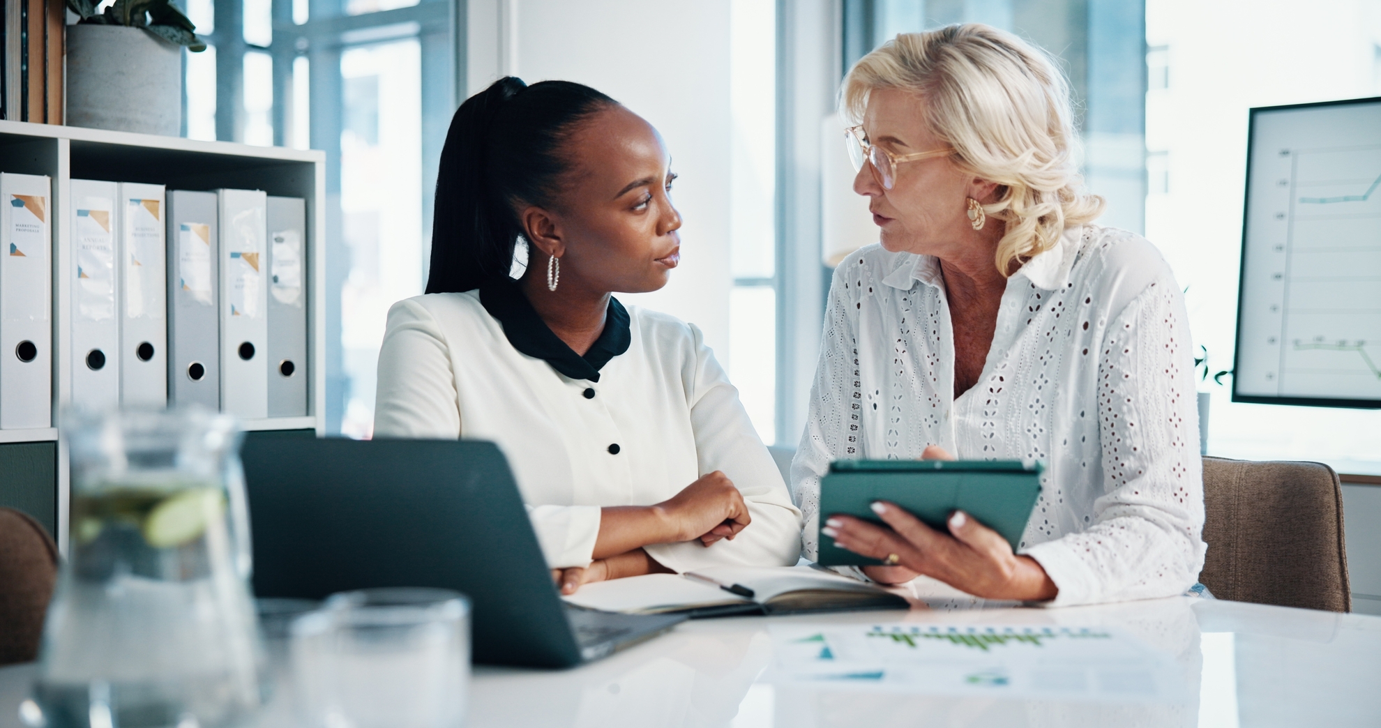 Dos contadores analizando información financiera en una reunión, revisando datos en una tablet y computador en un entorno de oficina.